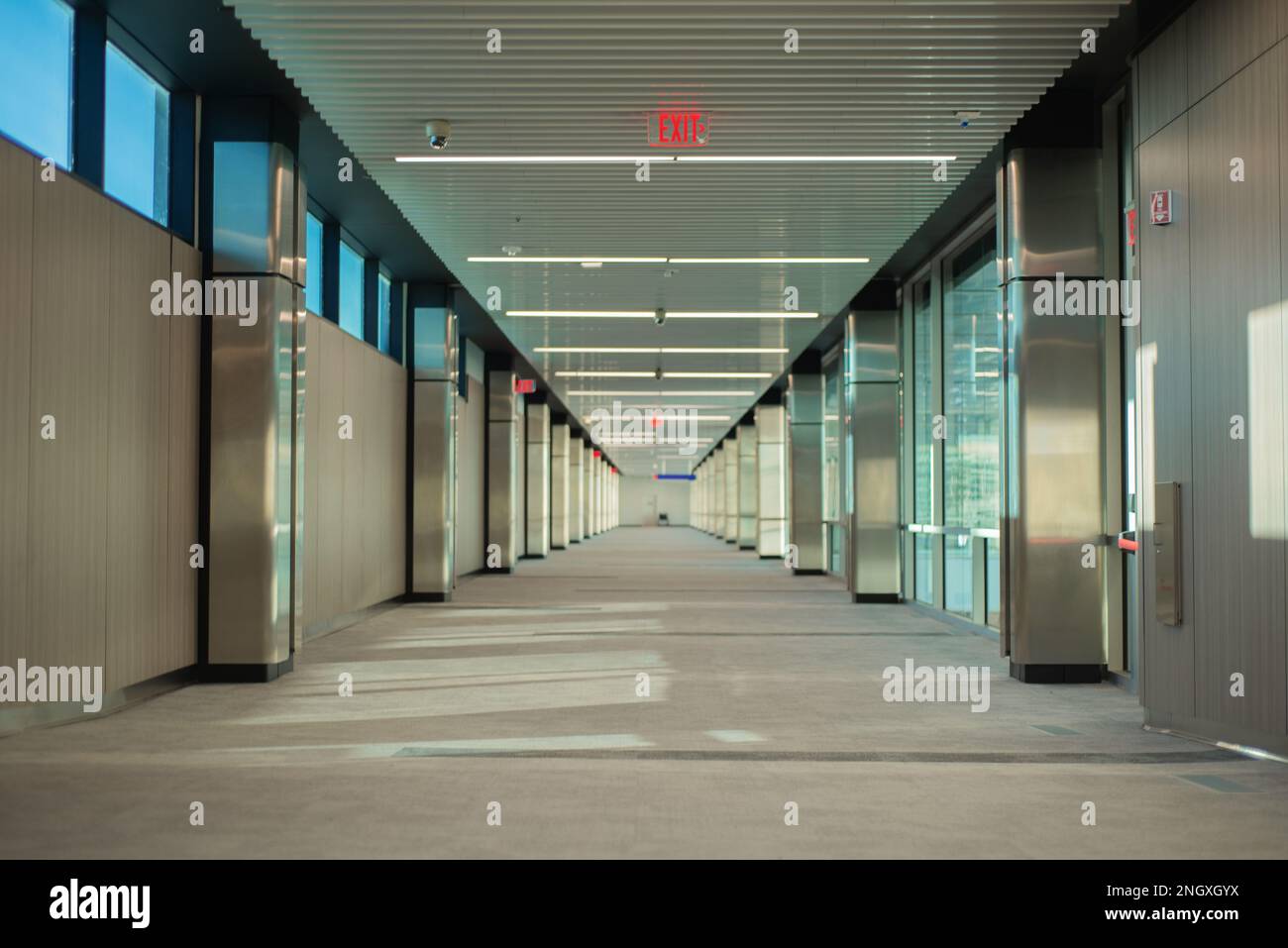 corridor of empty building with exit signs lit in red and windows on one side, no people Stock Photo