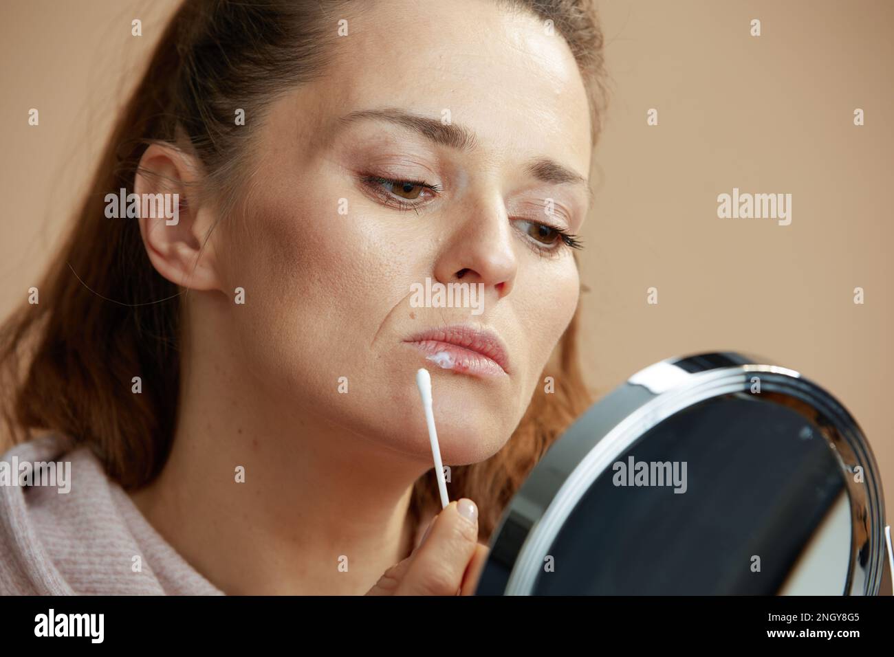 female with herpes on lips and mirror applying ointment using cotton swab isolated on beige