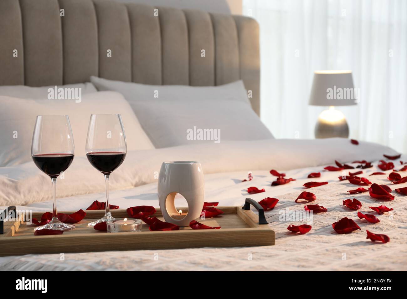 Tray with glasses of red wine, candles and rose petals on bed in room