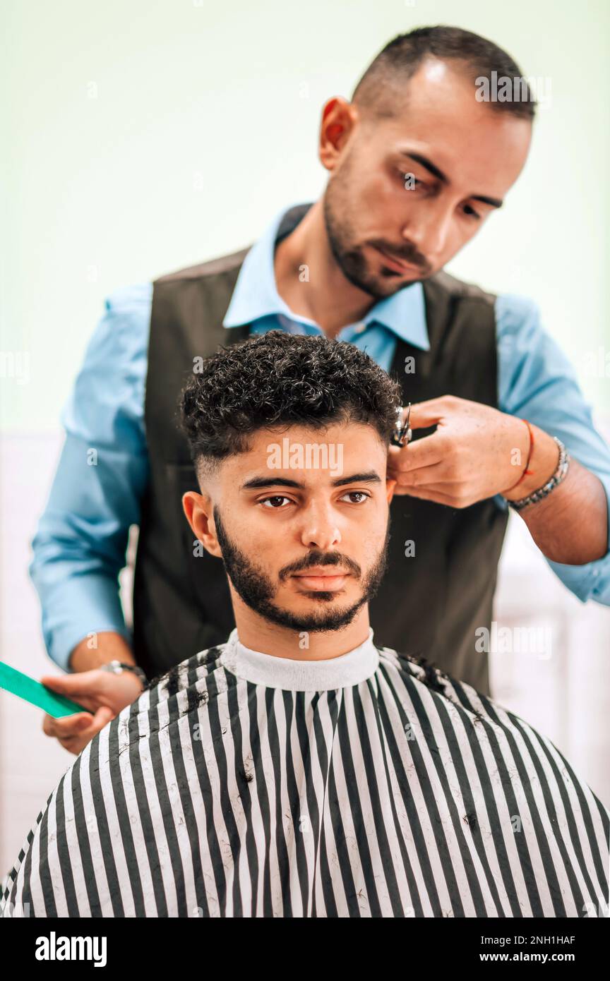 Serious bearded male client with dark curly hair in striped cape sitting at barbershop while barber making haircut Stock Photo