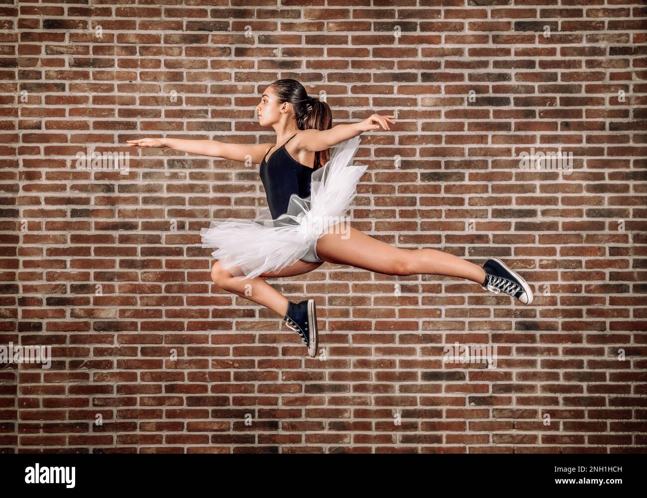 Side view full body of young ballerina in white tutu and black sneakers jumping against brick wall Stock Photo