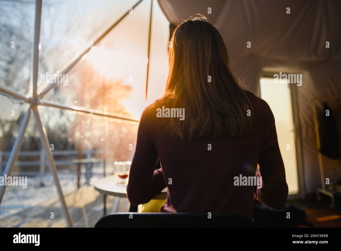 View from the back of a girl lit by the setting sun sitting at a table by the window. Close-up, a girl with a cup of coffee. Stock Photo