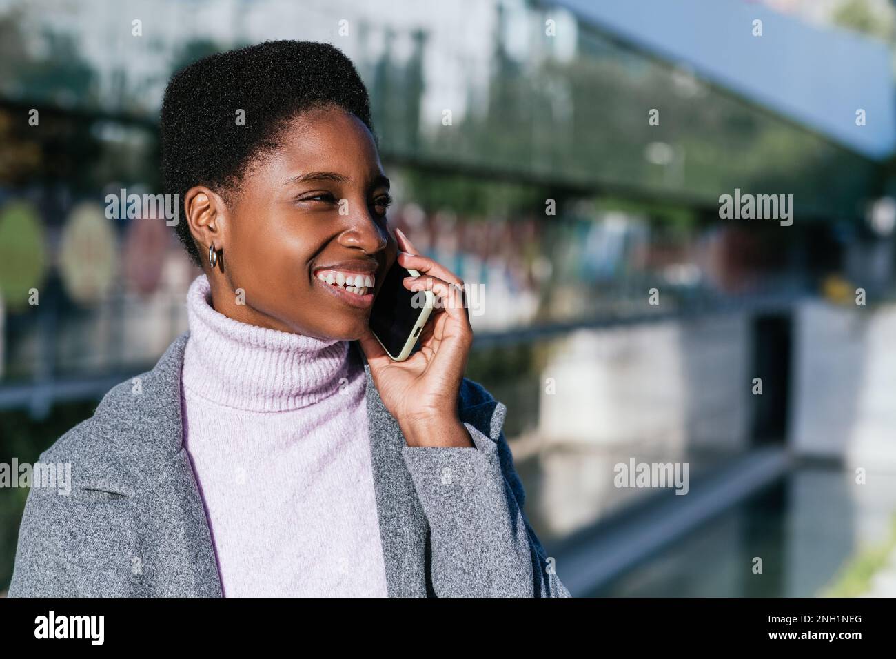 Cheerful African American female in stylish outerwear smiling and looking away while answering phone call on blurred background of city street Stock Photo