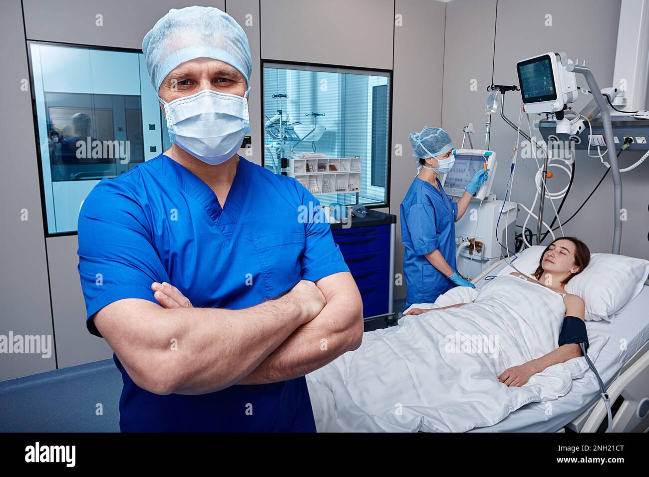Portrait of male nurse with arms crossed standing in hospital ward of ICU with unconscious female patient while caring for her Stock Photo
