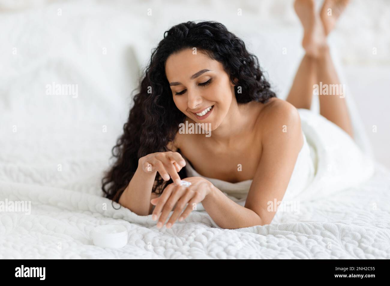 Happy millennial caucasian brunette lady with long hair lies on soft bed, applies cream on hands Stock Photo