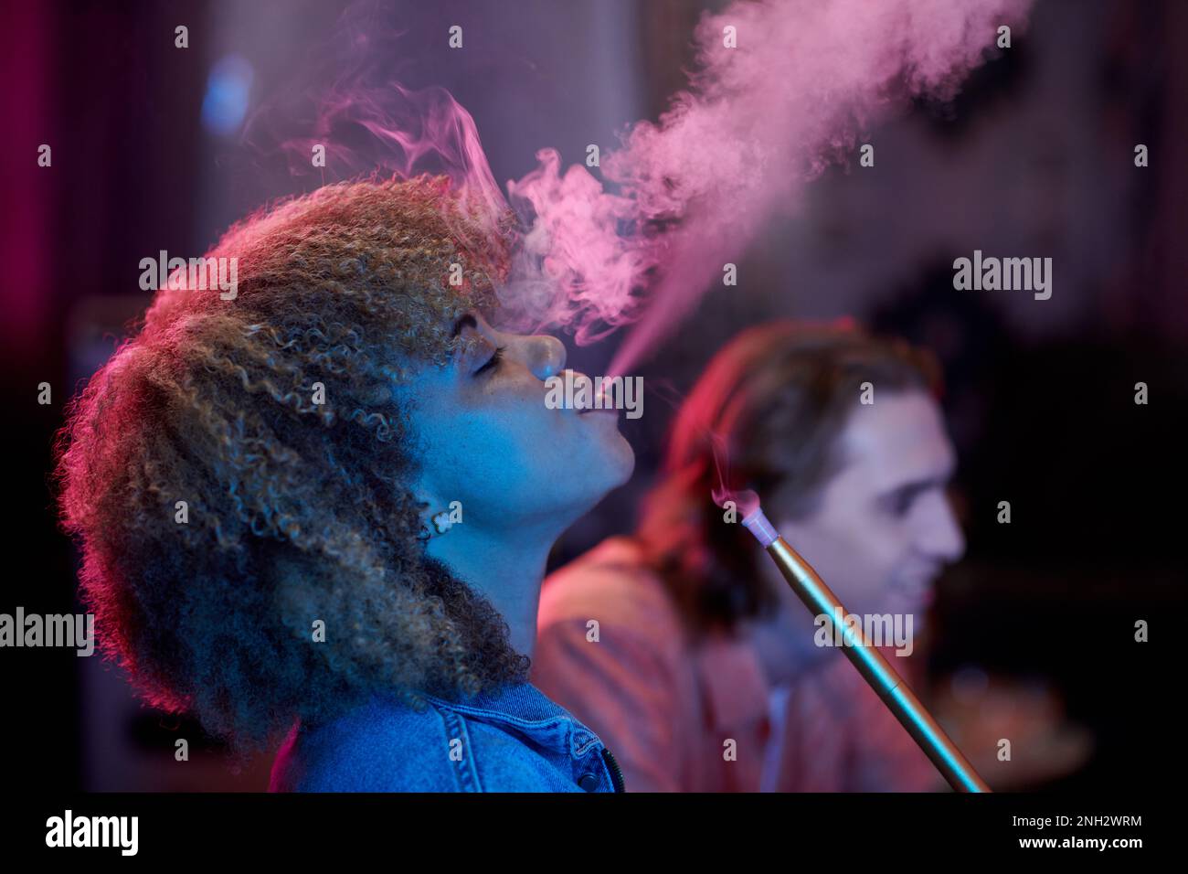 Side view of curly young woman exhaling smoke wneh enjoying hookah in night clubs Stock Photo