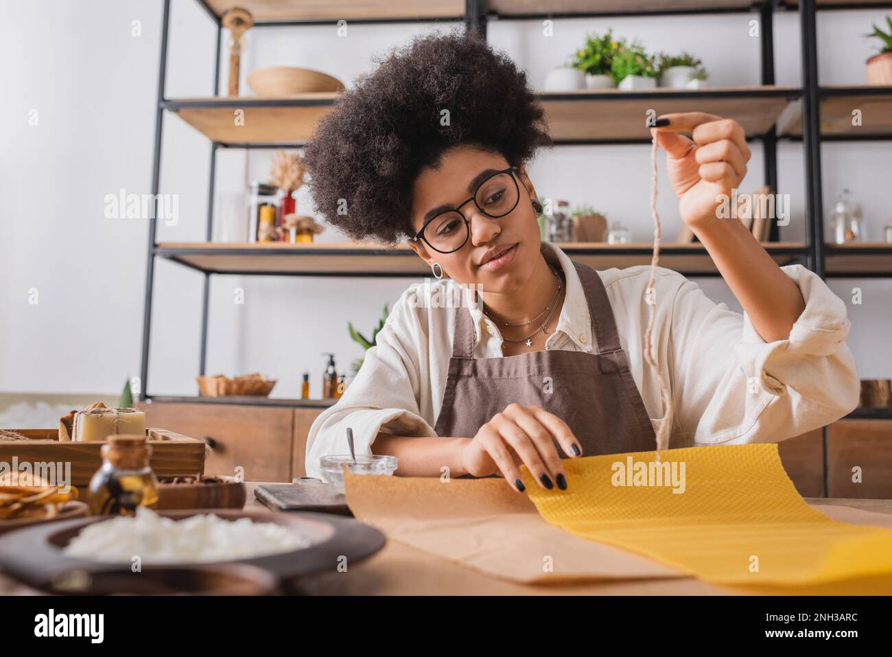 curly african american woman in eyeglasses and apron holding candle wick near wax sheet and natural ingredients on blurred foreground,stock image Stock Photo