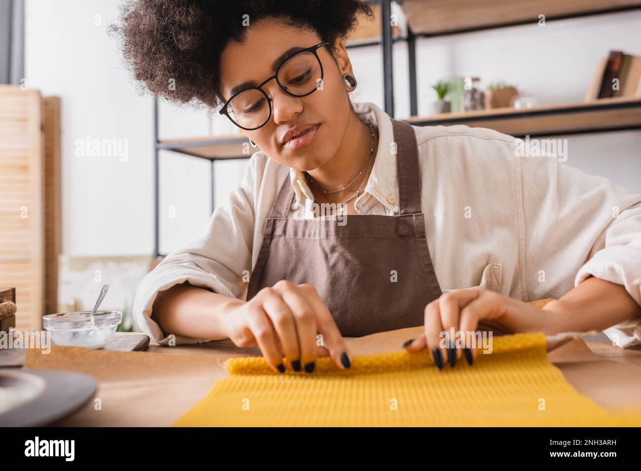 african american craftswoman in apron and eyeglasses rolling wax sheet on parchment while making candle in workshop,stock image Stock Photo