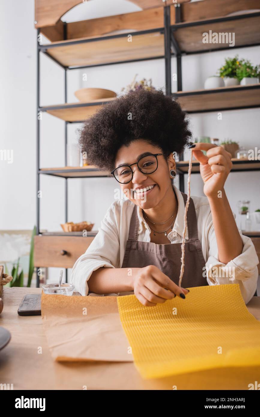 cheerful african american craftswoman in eyeglasses looking at camera while holding twine near wax sheet in workshop,stock image Stock Photo