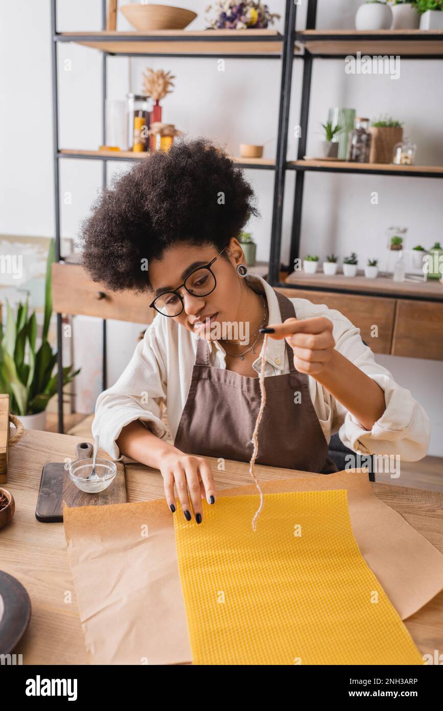 african american craftswoman in apron and eyeglasses holding candle wick near natural wax sheet and parchment on wooden table,stock image Stock Photo