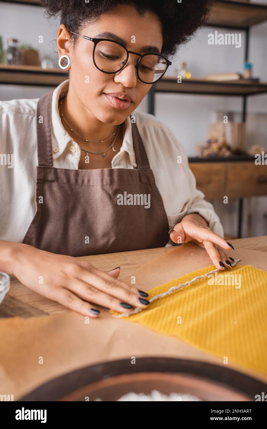 african american craftswoman in eyeglasses and apron holding candle wick near wax sheet on craft paper,stock image Stock Photo