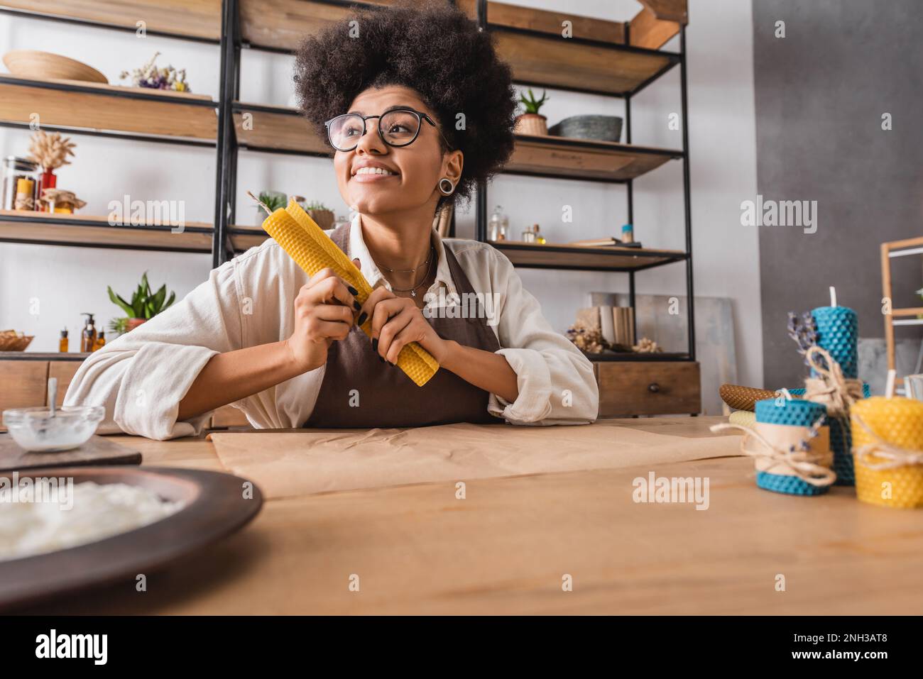 pleased african american woman in eyeglasses and apron holding rolled wax sheet near homemade candles in craft workshop,stock image Stock Photo