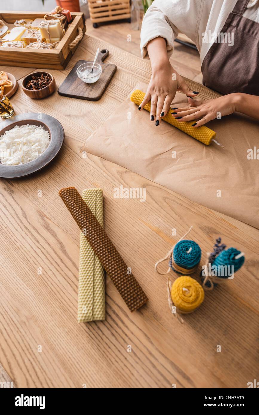 partial view of african american woman rolling wax sheet on parchment near homemade candles on wooden table,stock image Stock Photo