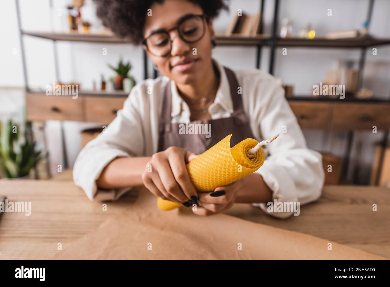 selective focus of handmade candle in hands of positive african american woman in eyeglasses and apron on blurred background,stock image Stock Photo