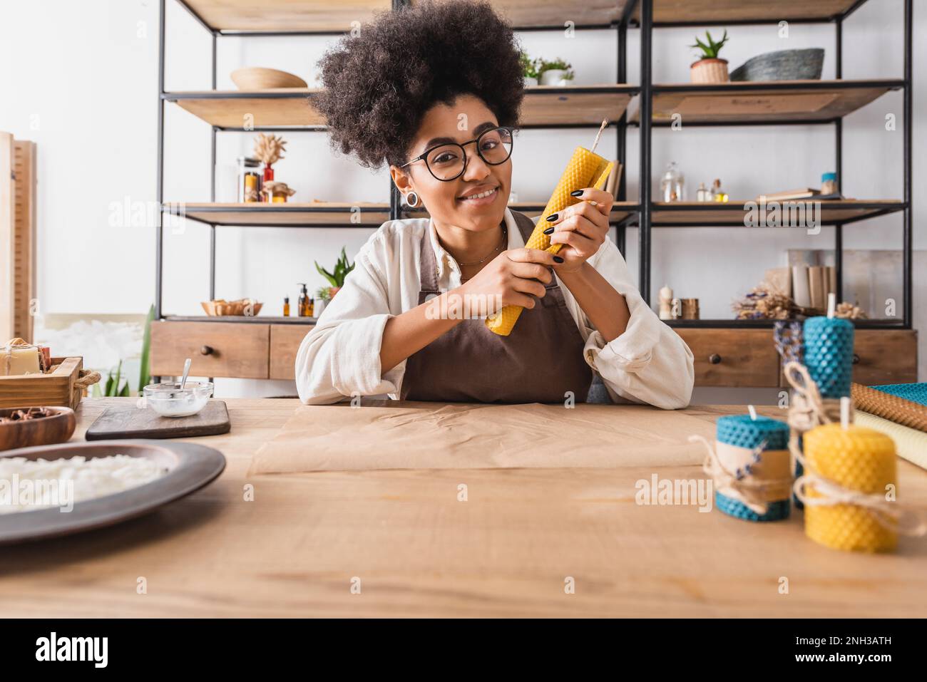 cheerful african american woman in eyeglasses looking at camera while sitting with handmade candle near parchment in workshop,stock image Stock Photo