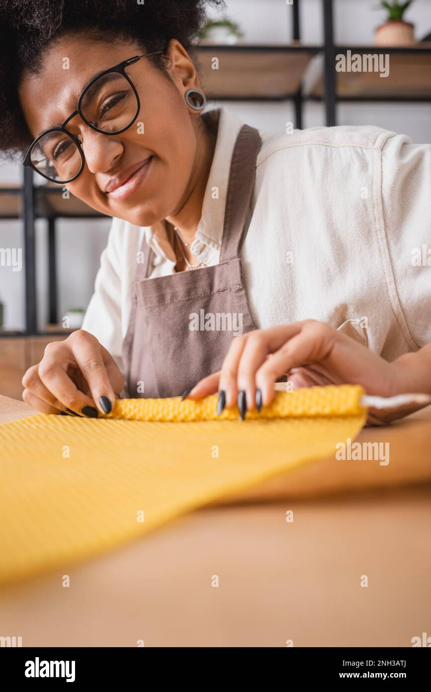 cheerful african american craftswoman in eyeglasses and apron smiling at camera while making candle in workshop,stock image Stock Photo