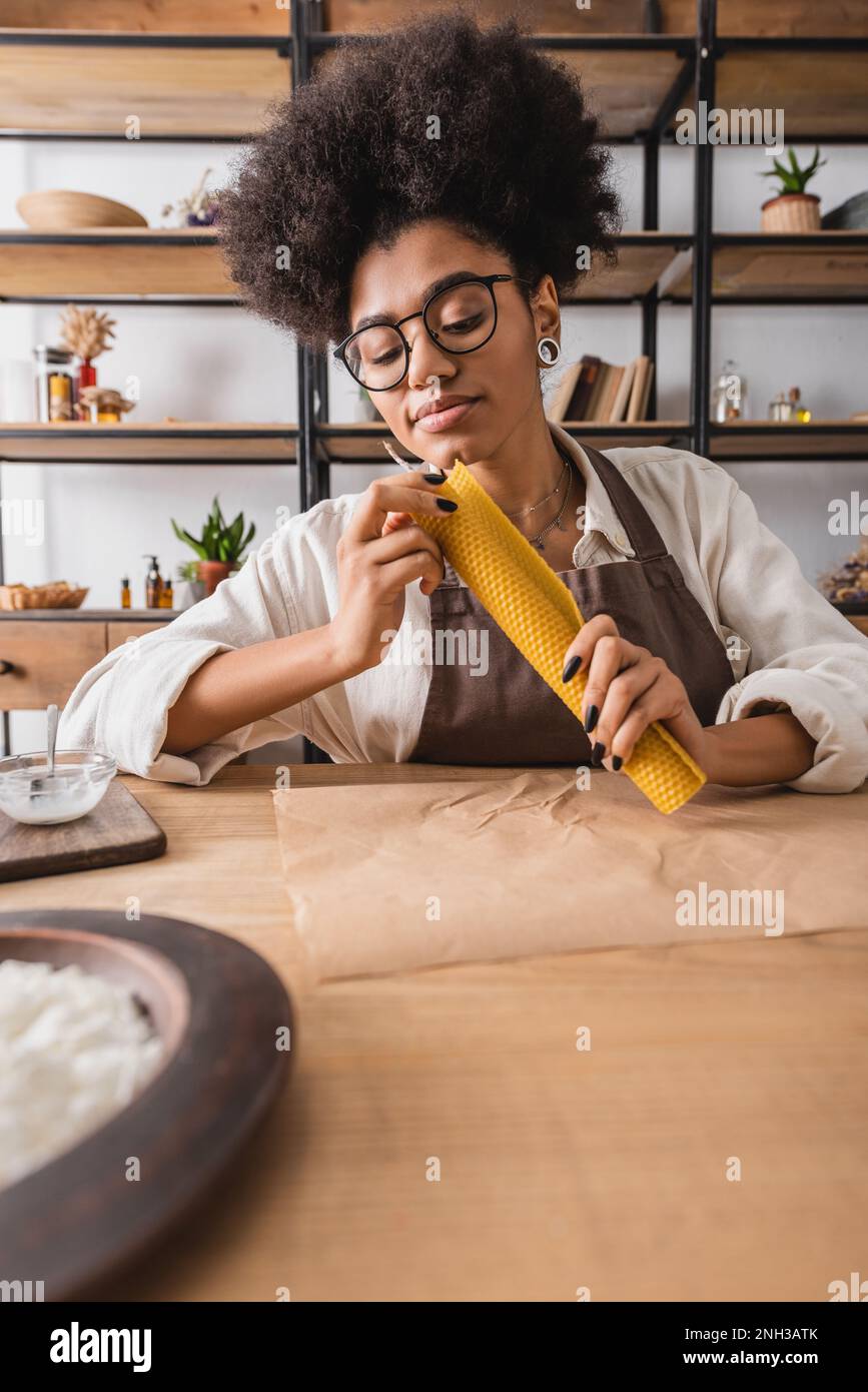 curly african american craftswoman in apron and eyeglasses making natural handle near parchment on table in workshop,stock image Stock Photo