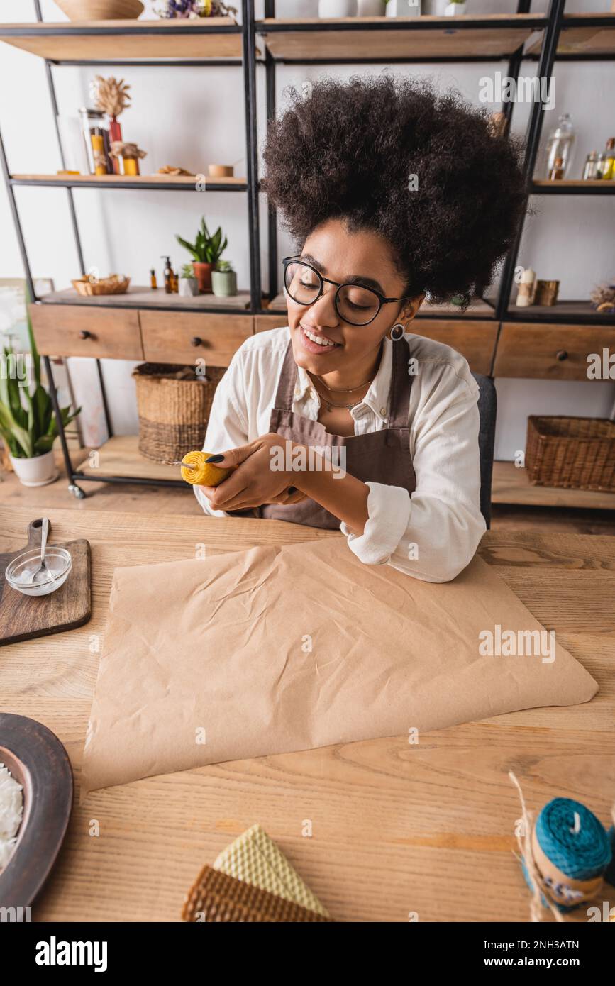 smiling african american craftswoman in apron and eyeglasses making natural candle near craft paper in workshop,stock image Stock Photo