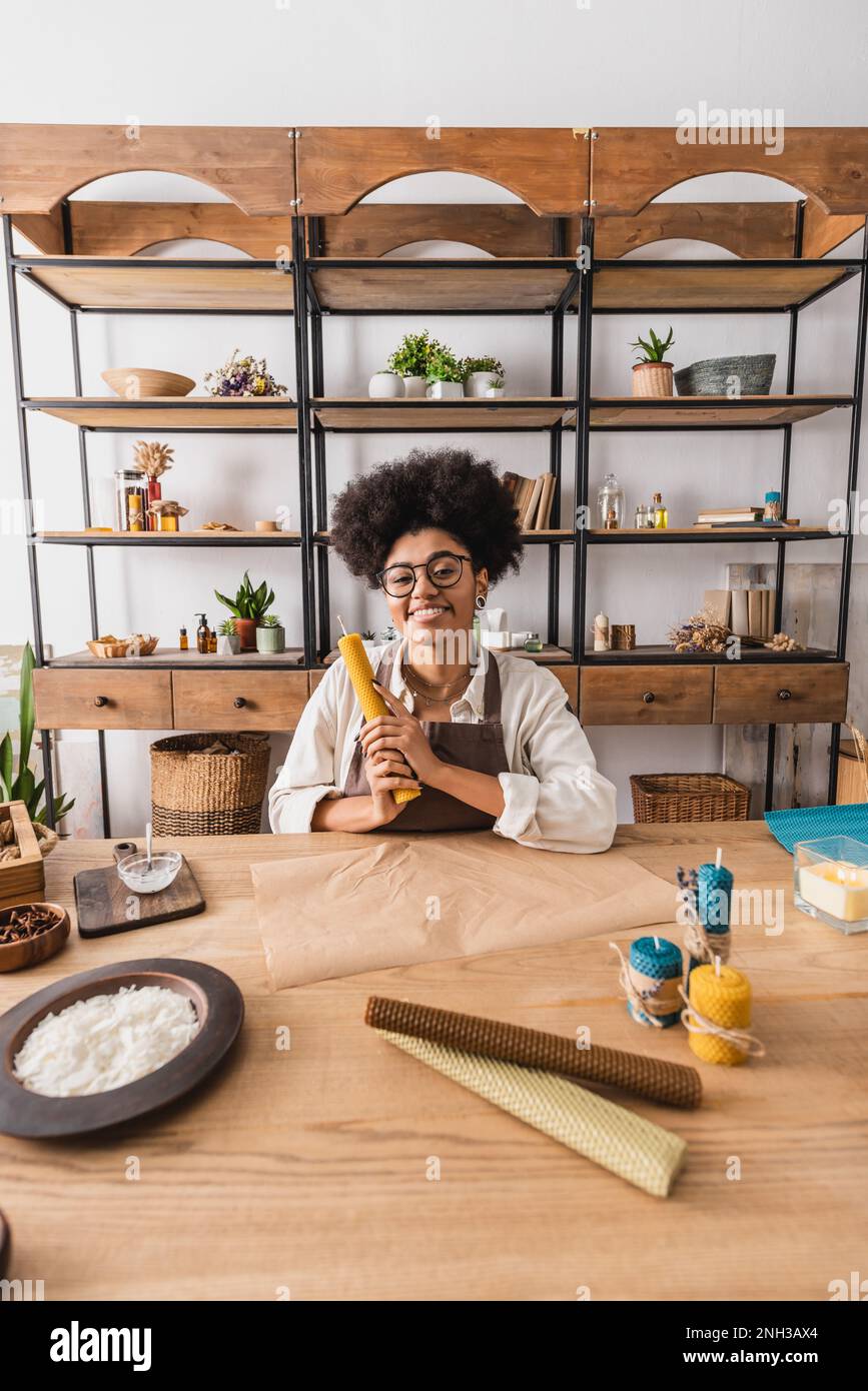 happy african american woman holding rolled wax sheet near parchment and handmade candles on table in craft workshop,stock image Stock Photo
