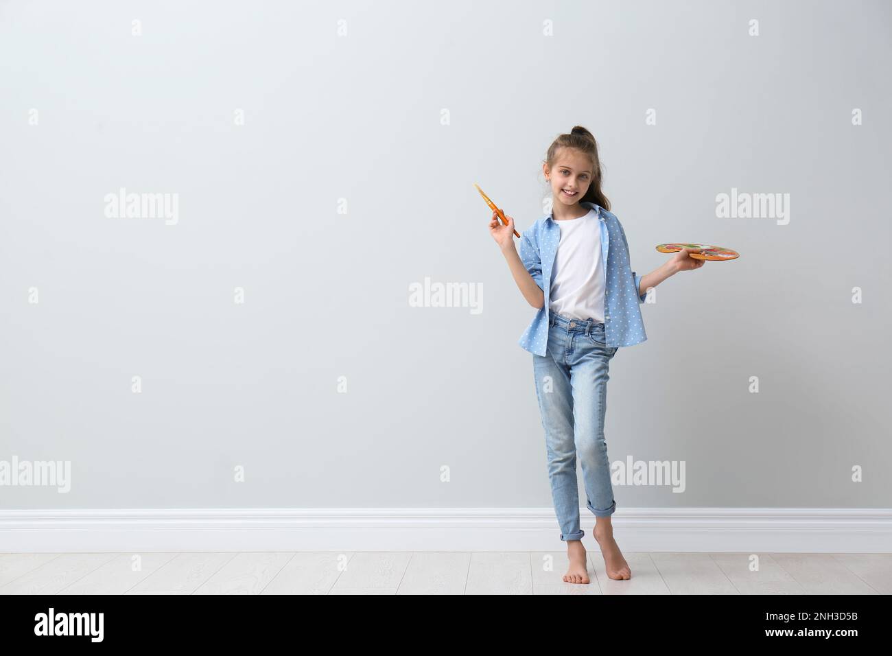 Little girl with brush and paint palette near light wall indoors. Space for text Stock Photo