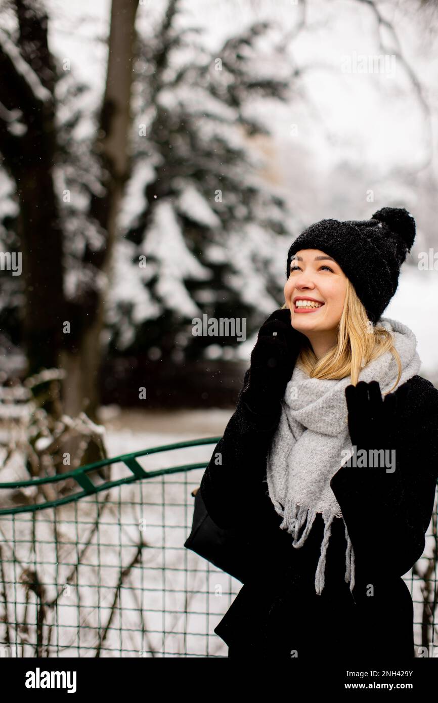 A young woman wearing warm winter clothes and a knit hat smiles happily as she stands in the snow nd using mobile phone Stock Photo