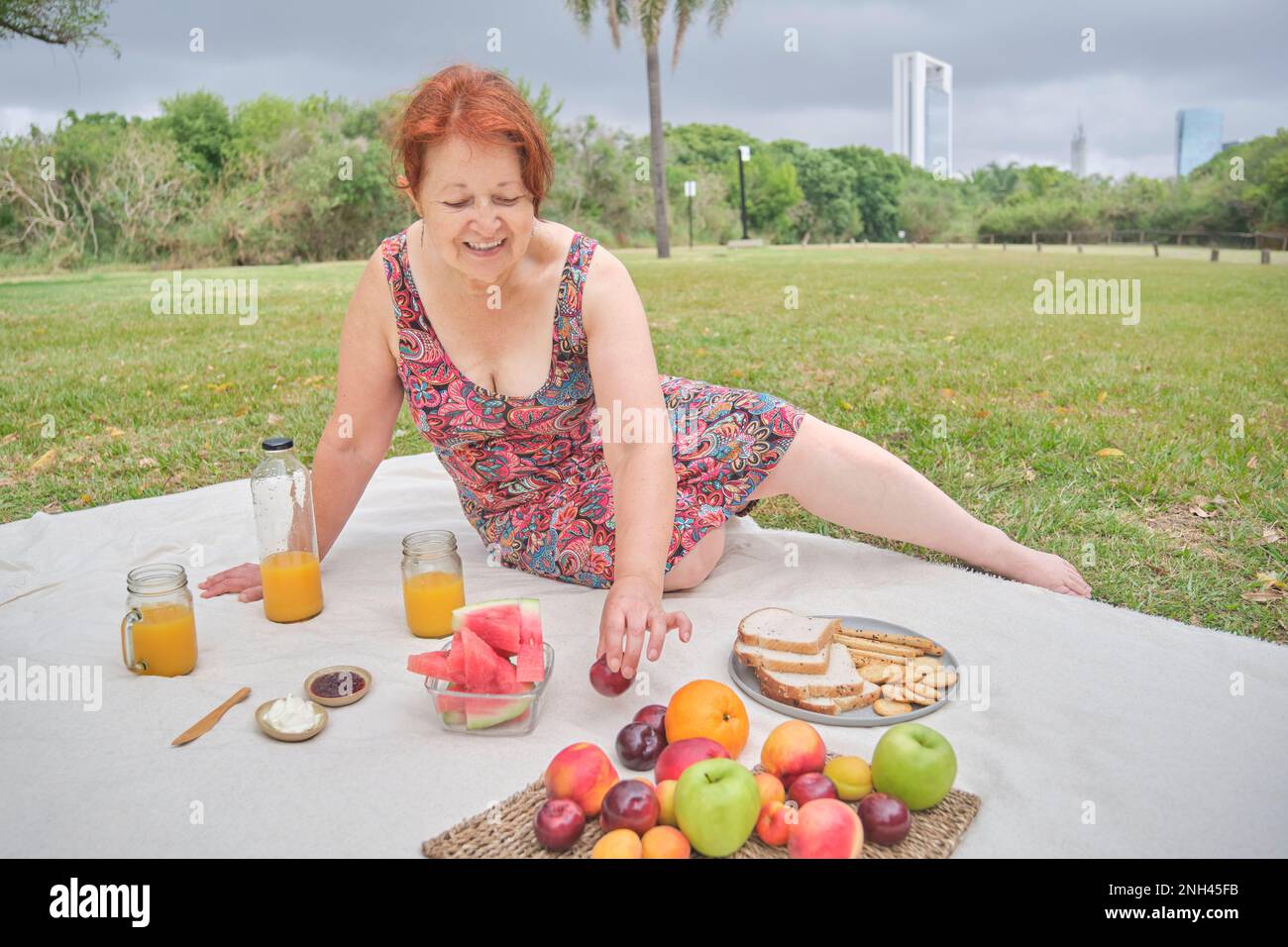 Happy mature hispanic woman enjoying a healthy breakfast picnic with whole grain bread and crackers and assorted fruits. Stock Photo