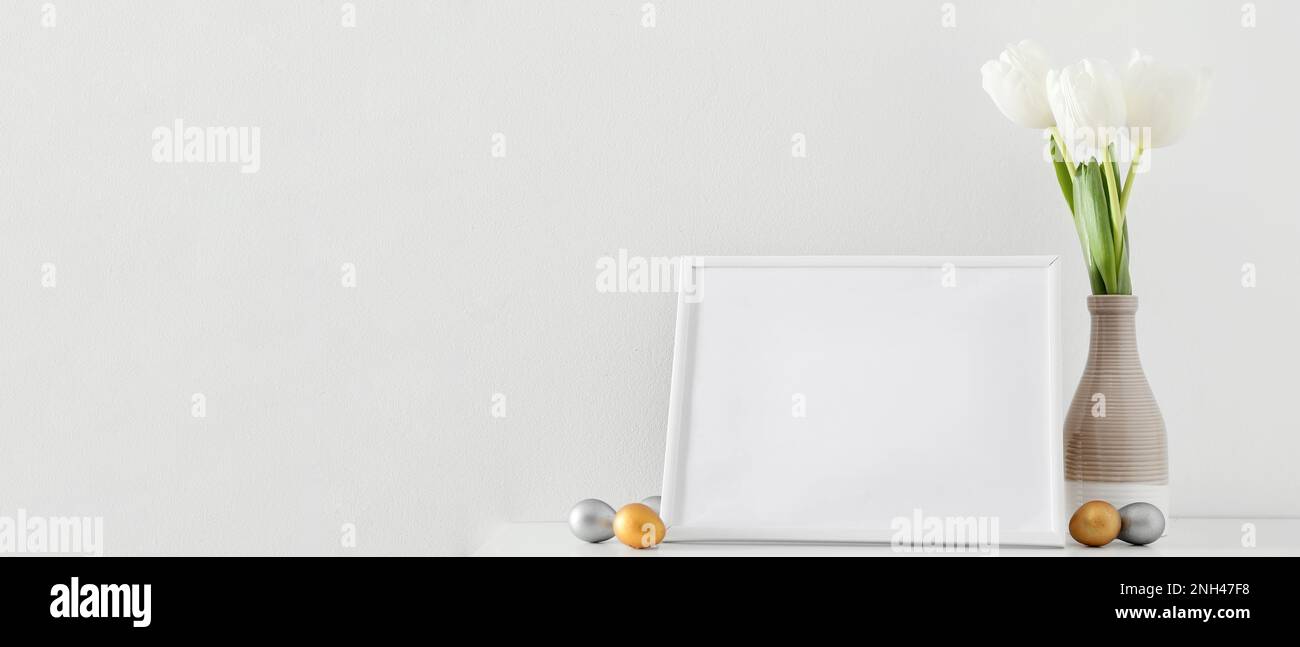 Blank photo frame, Easter eggs and vase with tulips on table near white ...