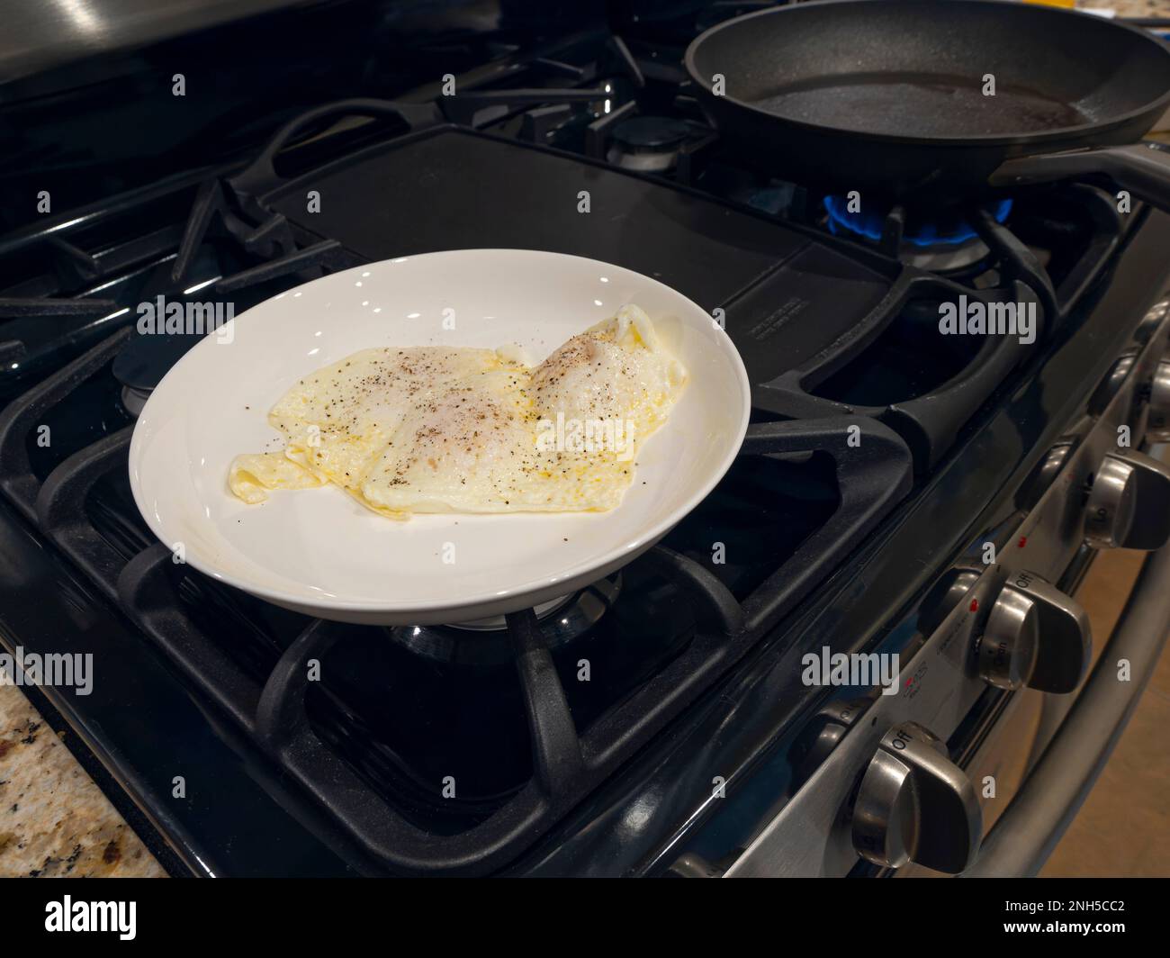 Fried eggs over medium, for breakfast, in a white dinner plate on a stovetop. Stock Photo