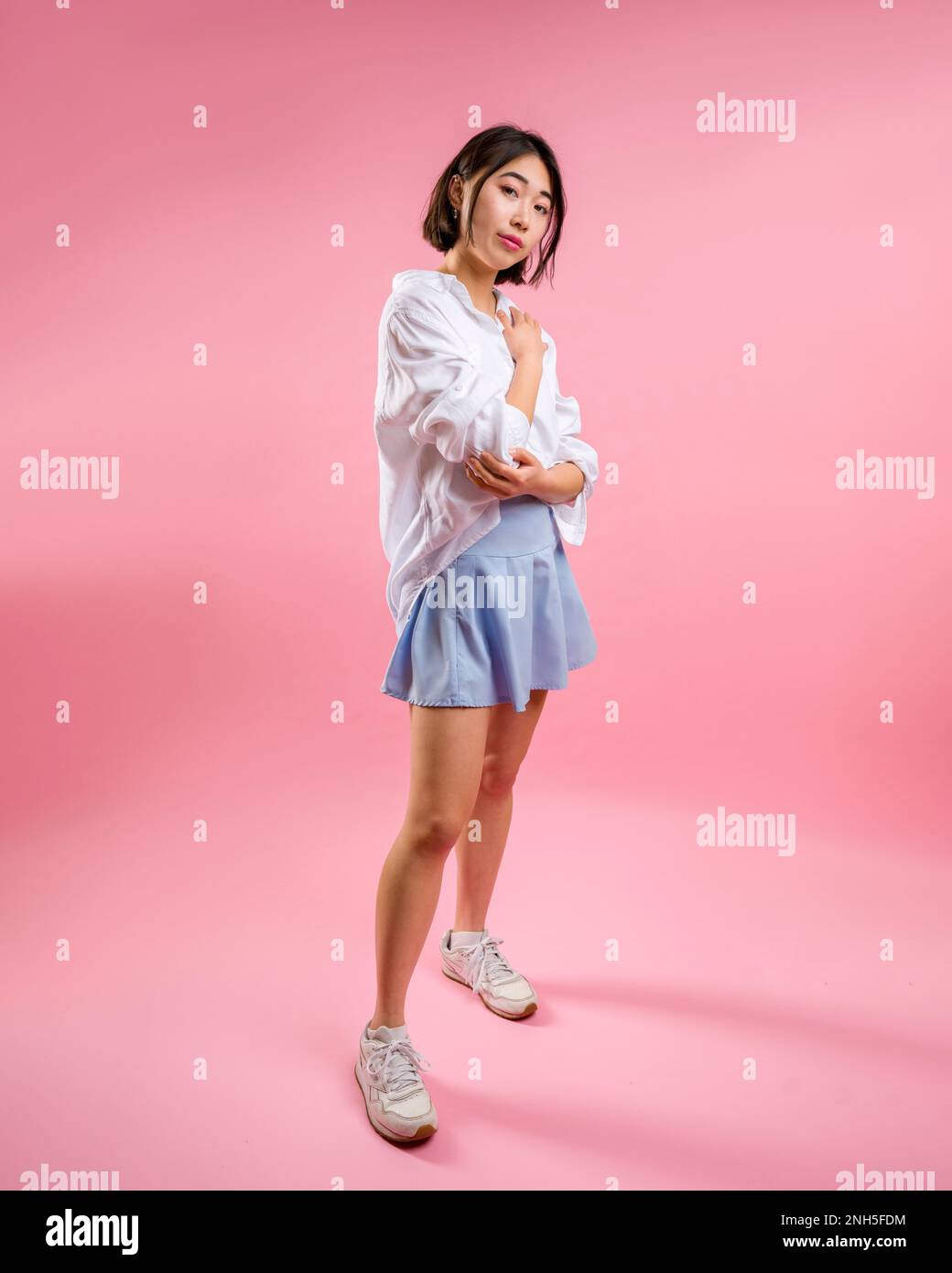 Full Body Asian Woman Short Blue Skirt White Blouse Pink Backdrop Standing with Arms Wrapped Around Waist and Chest Side View Stock Photo