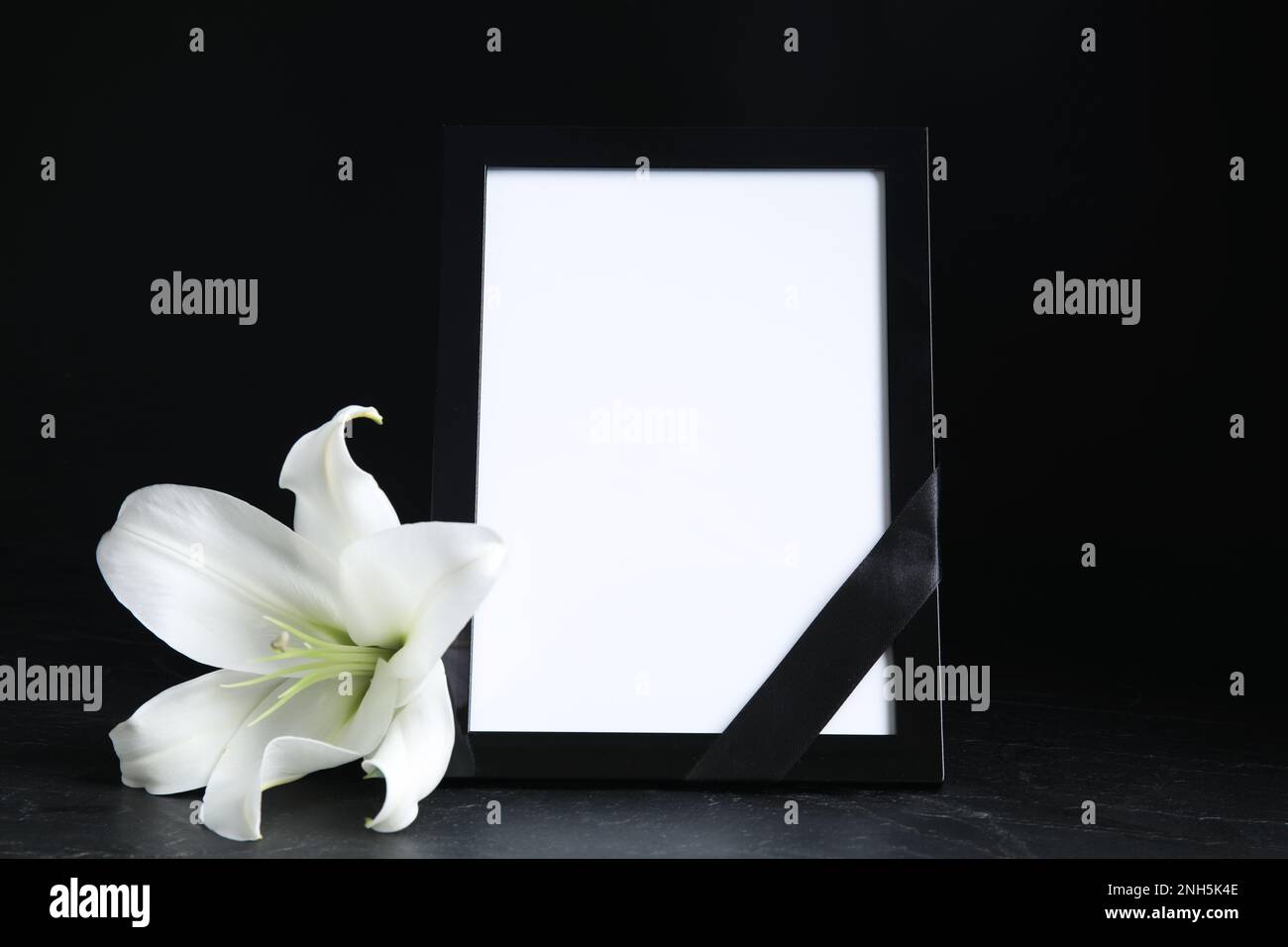 Funeral photo frame with ribbon and white lily on black table against