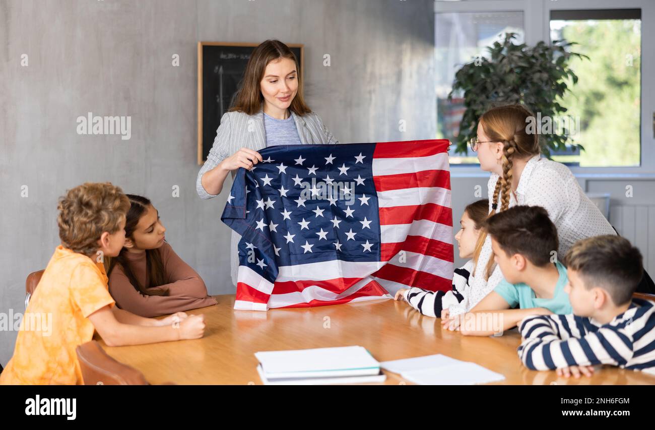 Smiling young woman teacher showing national flag of USA and telling preteens schoolchildren history of country during lesson in class Stock Photo