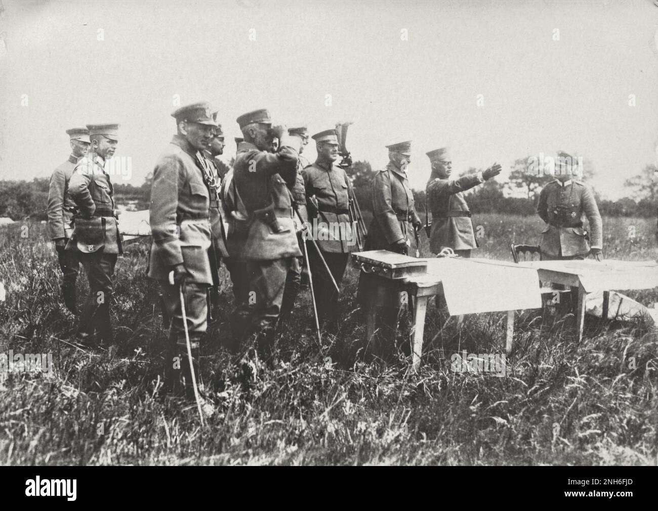 1914-1918. World War I. The German Emperor William II and his brother Henry of Prussia during a troop visit. The group with the emperor and high-ranking servicemen stands on a meadow with tall grass. In the foreground are two tables with maps and a chair. An officer points with his arm at something and everyone looks in this direction, the prince with the help of binoculars. Stock Photo