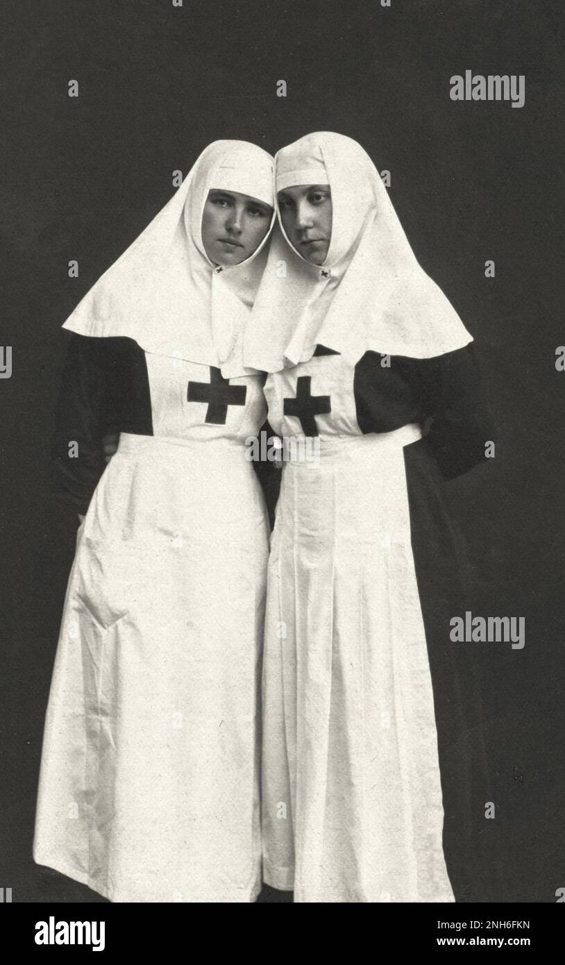 World War I. Two Latvian nurses standing side by side. 1915 Black and white postcard featuring a photographic portrait of two nurses standing next to each other with their heads touching and their hands behind their backs. Both wear long black dresses, white aprons and white nurse's veils that cover their hair. They each have a cross on their uniform's pinafore Stock Photo