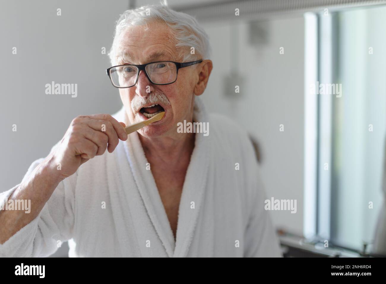 Senior man cleaning his teeth with wooden brush in bathroom, sustainable lifestyle. Stock Photo