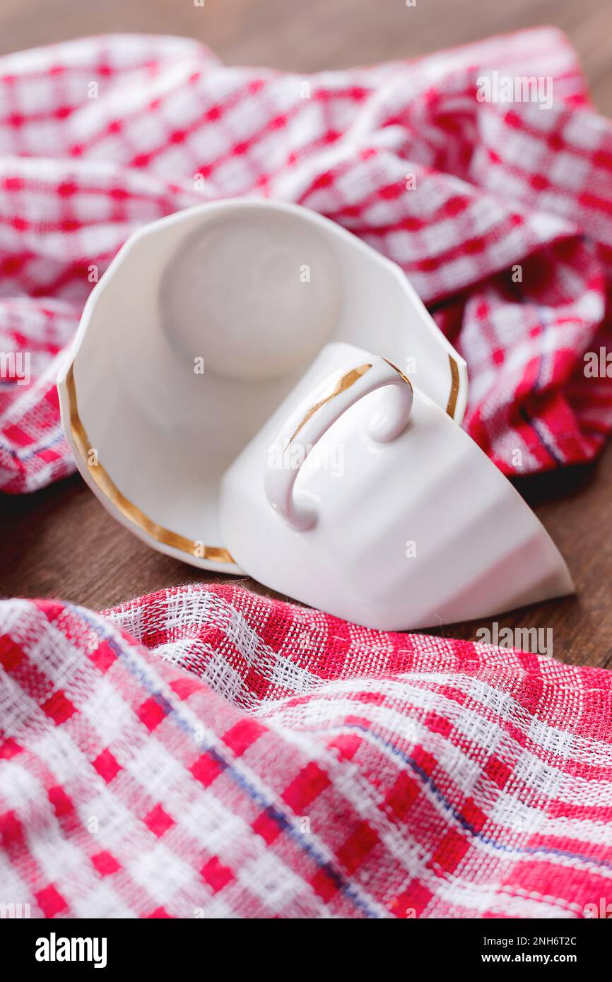 Broken white cup on wooden background with plaid red towel. Damaged mug with golden decoration. Stock Photo