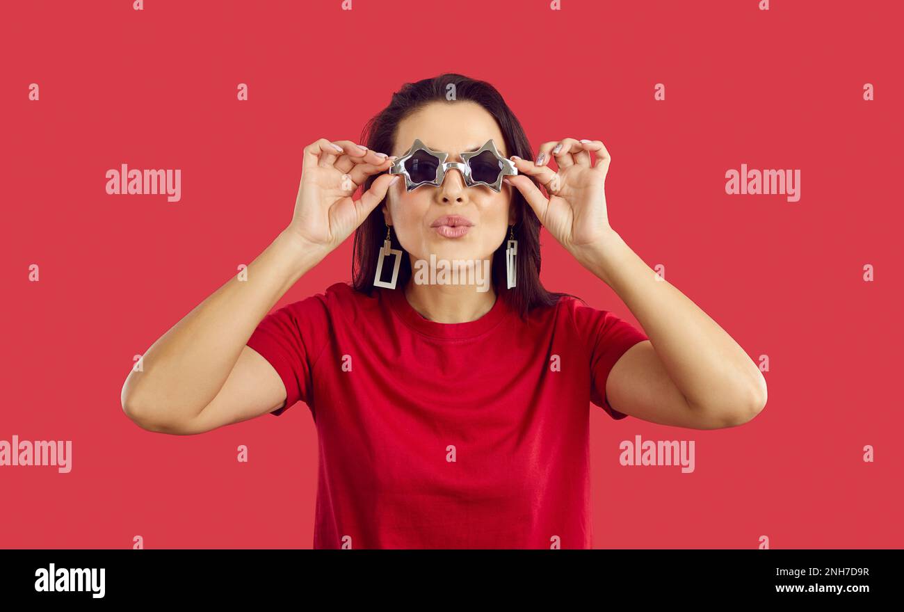 Cool and positive woman in star shape glasses makes an air kiss on vivid red background. Stock Photo
