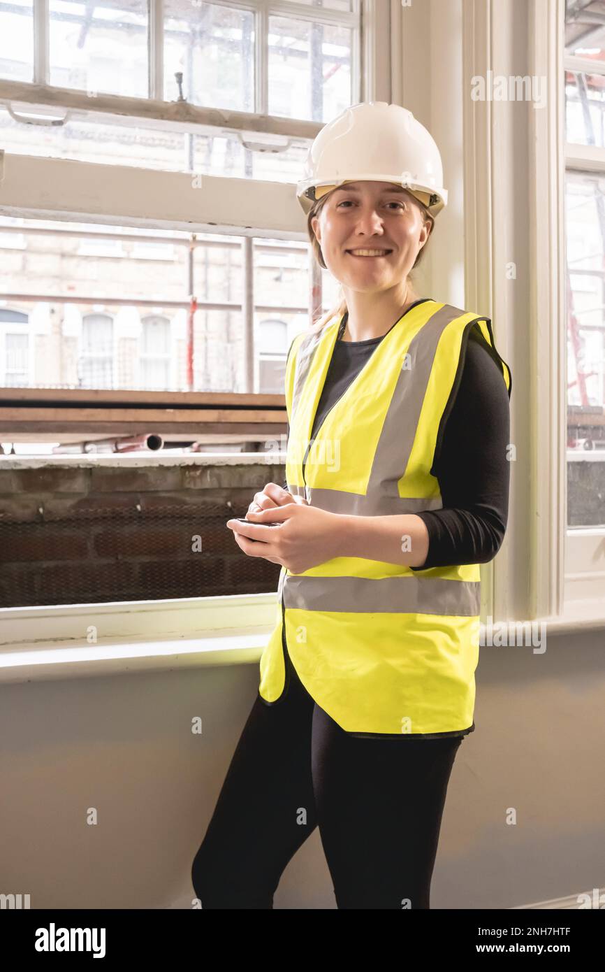 Vertical photo of a smiley chartered civil engineer woman checking budget against spend with a calculator in a construction site, hard hat, yellow per Stock Photo