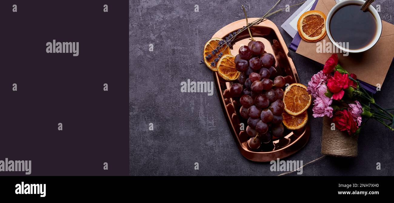 Aesthetic grapes on the golden tray with cup of coffee, dry oranges, books and flowers, copy space. Extra wide banner. Stock Photo
