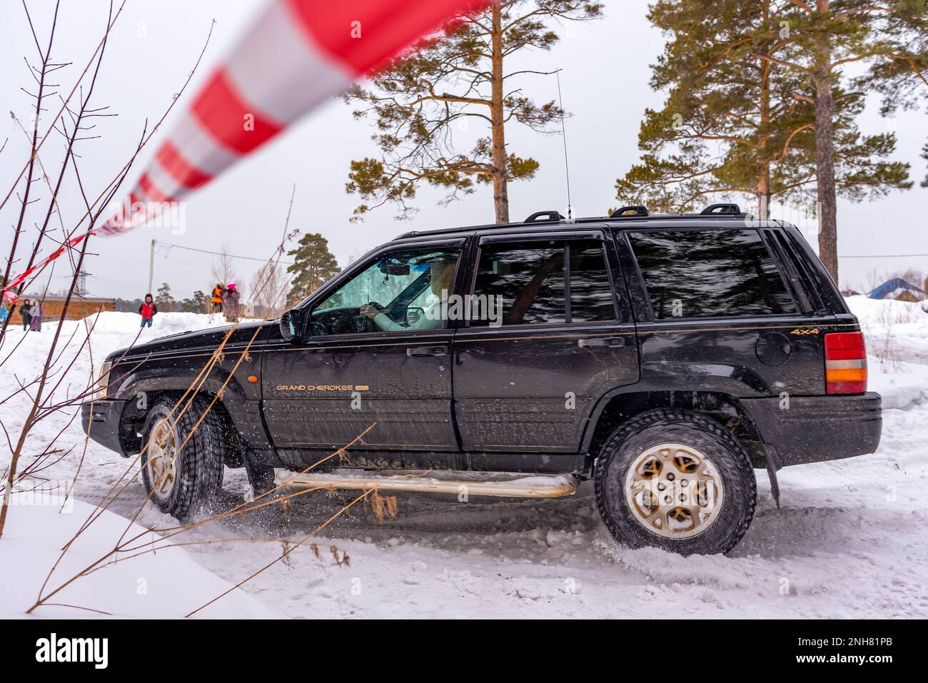 Offroad SUV 'Jeep Grand Cherokee' 4x4 black color rides in winter on snow in the forest among pines and striped tape with a woman driver. Stock Photo