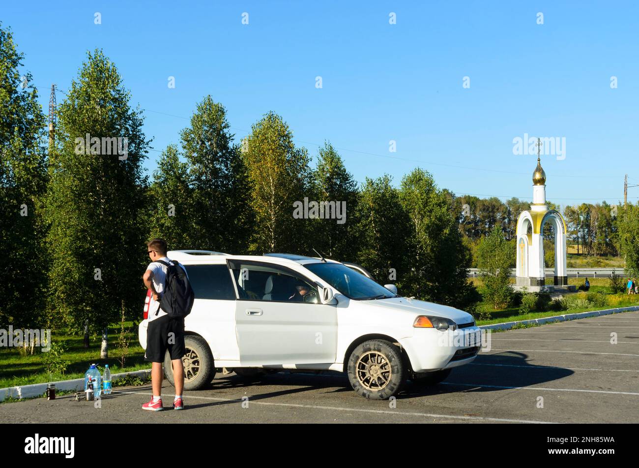 Male hiker with backpack in sneakers 'Nike' is the car 'Honda' next to the gas burner and the water bottles in front of a chapel and tourists Stock Photo