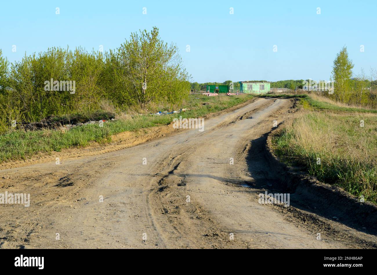 Dirt road with garbage in the grass to a small military building control checkpoint near the fence and the field with the forest in the summer. Stock Photo