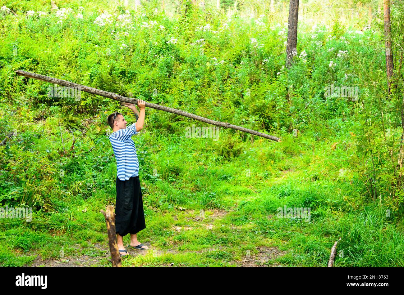 The man in the wide trousers yoga doing exercises turns the big stick in the woods on the grass. Stock Photo