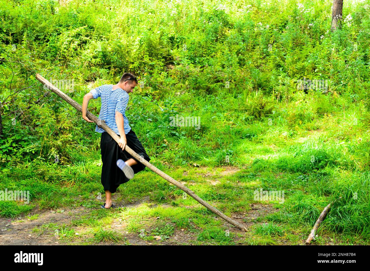 The man in the wide trousers yoga doing exercises turns the big stick in the woods on the grass. Stock Photo