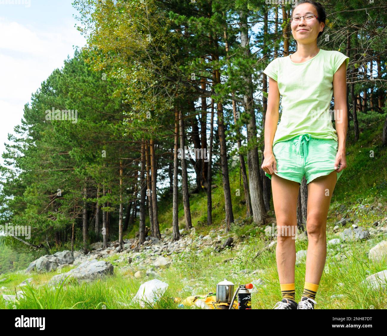 Happy Yakut girl in glasses stands on the banks of the river on the stones near the forest, holding his shorts and smiling at a picnic in the Altai Stock Photo