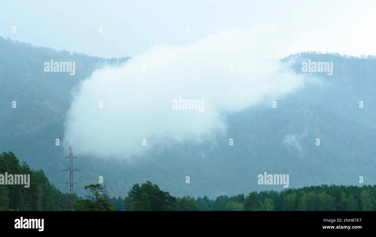 Strange single rainy Cumulus cloud floats in the fog mountains in the background above the trees of the forest next to the utility pole in the Altai. Stock Photo