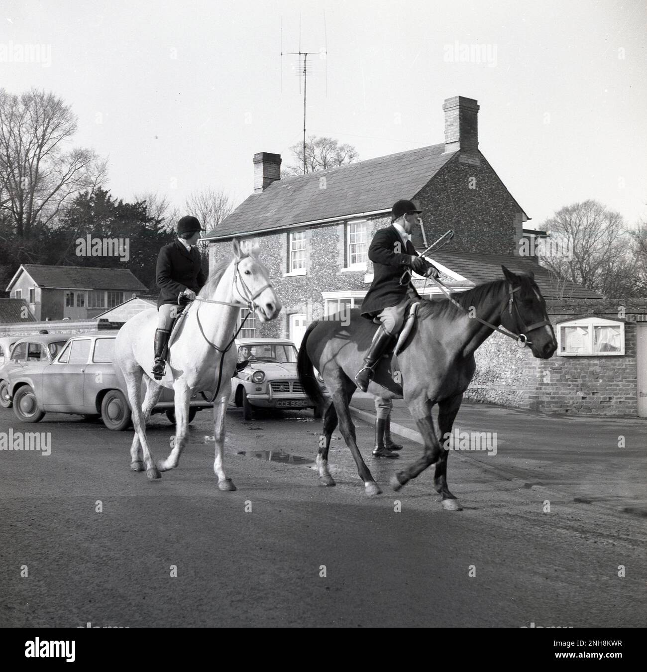 1965, historical, Thurlow Hunt, male and female hunt masters on their horses on a country lane, Thurlow, England, UK. The orgins of the Thurlow Hunt date back to the 18th century century, with the hunt taking place on arable and grassland that lies in both Suffolk and Cambridge. Cars of the era, including a Ford Anglia parked on the side of the road. Stock Photo