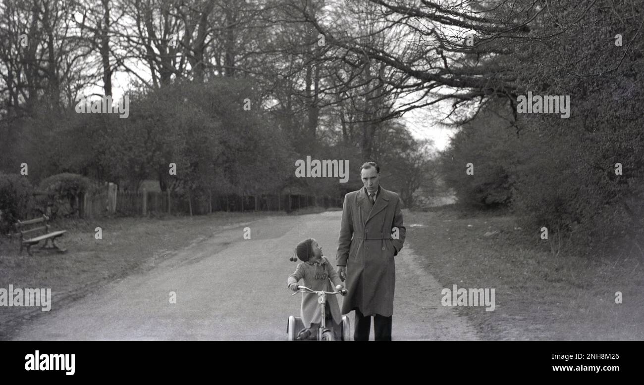 1950s, historical, a little girl in coat and woollen bobble hat riding a steel framed tricycle along a quiet, semi-rural country lane, with her father, who is wearing a trench coat, walking beside her, England, UK. Stock Photo