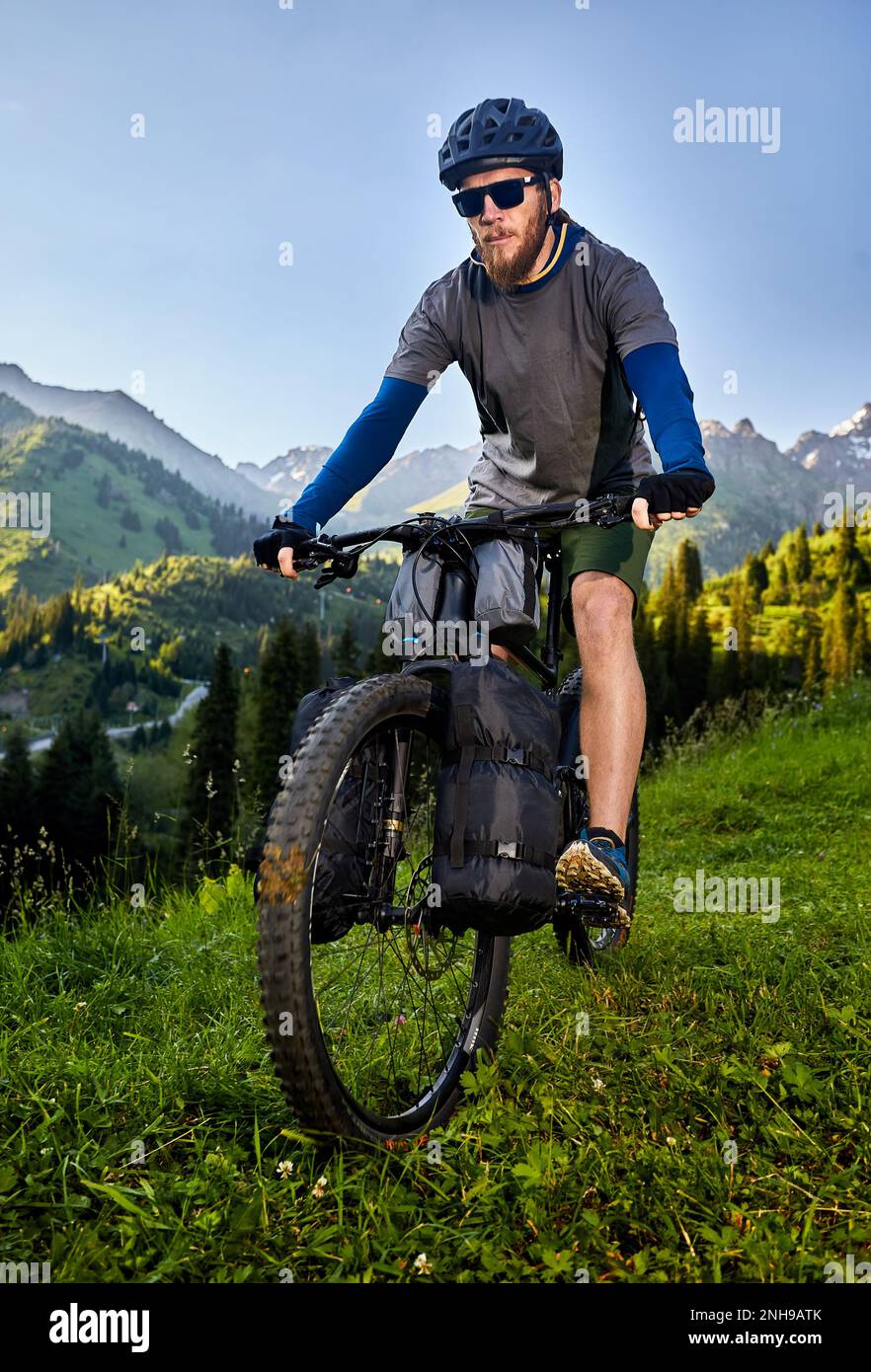 Man with dreadlocks and beard ride mountain bike with tourist bags at the green hill forest in beautiful mountain valley at sunset in Almaty, Kazakhst Stock Photo