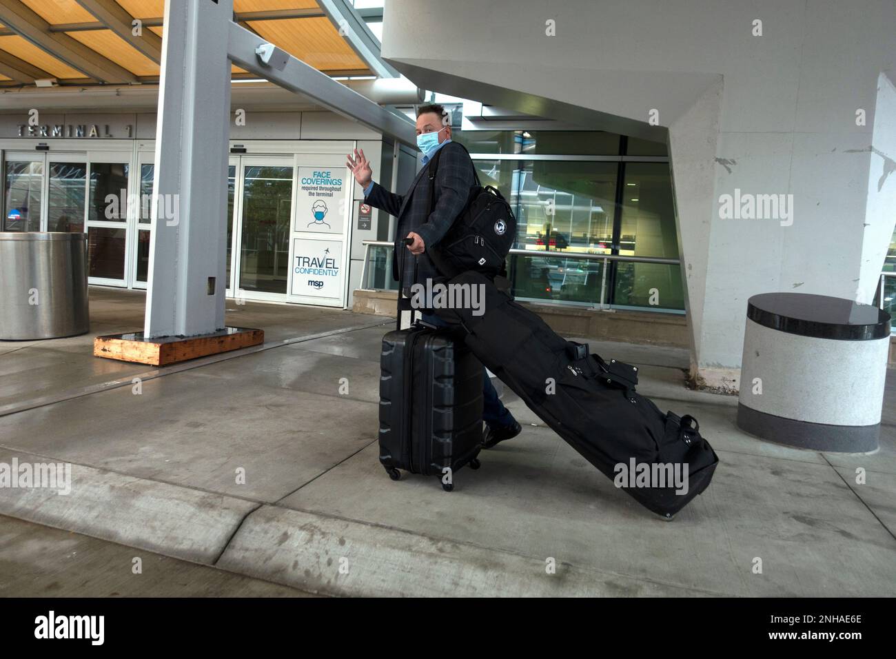 Man wearing a masked for Covid protection with golf clubs and carry-on luggage waving at airport traveling by airplane. Minneapolis Minnesota MN USA Stock Photo
