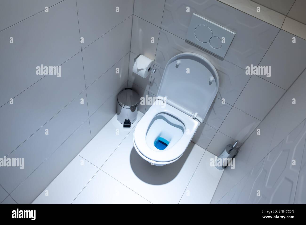 View of the white toilet bowl in the toilet. Bathroom finished with white glaze tiles and white terracotta. Trash can and brush in the background Stock Photo